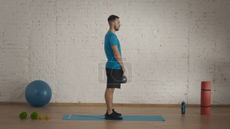 Photo for Side view. Athletic male fitness instructor doing exercises at the home studio for online classes. Man in sportswear doing squats with kettlebell. Healthcare concept. - Royalty Free Image