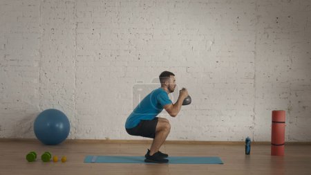 Photo for Athletic male fitness instructor doing exercises at the home studio for online classes. Man in sportswear doing low squats with kettlebell. Healthcare concept. - Royalty Free Image