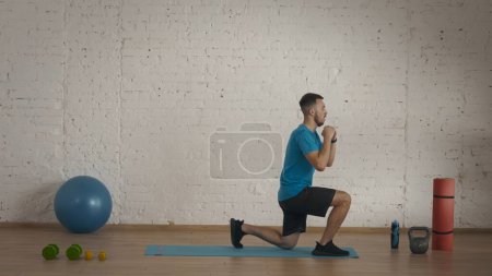 Photo for Personal sport trainings at home. Athletic male fitness coach doing exercises at the home studio for online classes. Man in sportswear doing jumping lunges exercise. - Royalty Free Image