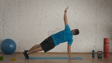 Foto de Entrenador de fitness masculino haciendo ejercicios en el estudio en casa para clases en línea. Hombre en ropa deportiva haciendo ejercicio de tablón activo con estiramiento. - Imagen libre de derechos