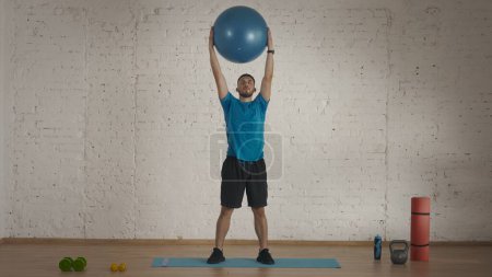 Photo for Sport lesson indoors. Athletic male fitness coach doing exercises at the home studio for online classes. Man in sportswear doing body bends exercise with a fitball. - Royalty Free Image