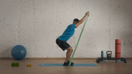 Photo for Sports indoors. Athletic male fitness coach doing exercises at the home studio for online classes. Man in sportswear doing half squat rubber band exercise. - Royalty Free Image