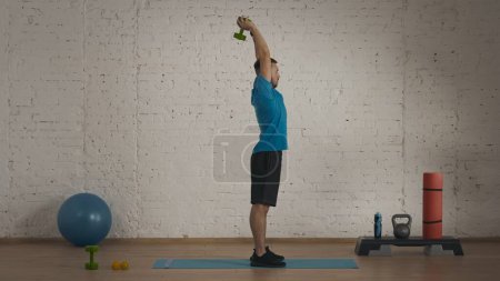 Photo for Sport indoors. Athletic man coach doing exercises at the home studio for online classes. Man in sportswear doing fitness triceps exercise. Side view. - Royalty Free Image