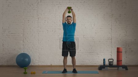 Photo for Athletic fitness coach doing exercise at the home studio for online classes. Man in sportswear doing triceps exercise with dumbbell. Bodycare concept. - Royalty Free Image