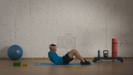 Photo for Sport indoors. Athletic fitness coach doing exercises at the home studio for online classes. Man in sportswear doing classic abs crunches exercise. - Royalty Free Image