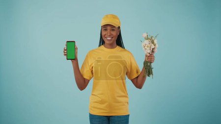 Téléchargez les photos : Concept de service de messagerie. Portrait d'une livreuse en bonnet jaune et tshirt tenant bouquet de fleurs et smartphone, souriant à l'écran. Espace publicitaire, maquette de l'espace de travail. - en image libre de droit