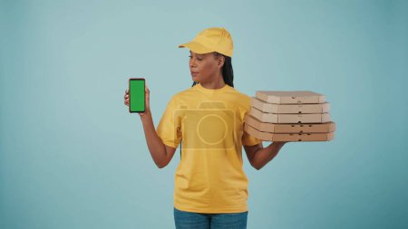 Photo for Portrait of a delivery woman in yellow cap and tshirt holding pizza boxes and smartphone, looking at the screen. Isolated on blue background. Advertising area, workspace mockup. - Royalty Free Image