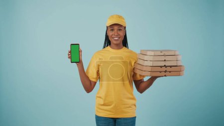 Photo for Portrait of a delivery woman in yellow cap and tshirt holding pizza boxes and smartphone, smiling at the camera. Isolated on blue background. Advertising area, workspace mockup. - Royalty Free Image