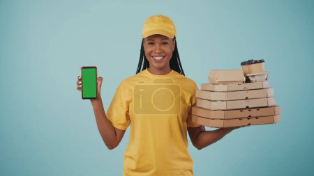 Téléchargez les photos : Portrait d'une livreuse en bonnet jaune et t-shirt tenant des boîtes à pizza, du café et un smartphone, souriant à la caméra. Isolé sur fond bleu. Espace publicitaire, maquette de l'espace de travail. - en image libre de droit