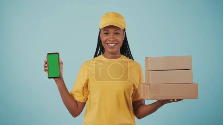 Photo for Portrait of a delivery woman in yellow cap and tshirt holding stack of postal boxes and smartphone, smiling. Isolated on blue background. Advertising area, workspace mockup. - Royalty Free Image