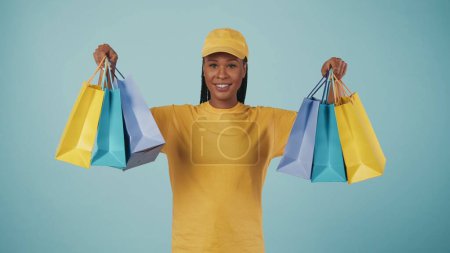 Téléchargez les photos : Portrait d'une livreuse en bonnet et t-shirt jaune tenant des sacs-cadeaux colorés dans les mains et souriant à la caméra. Isolé sur fond bleu. - en image libre de droit