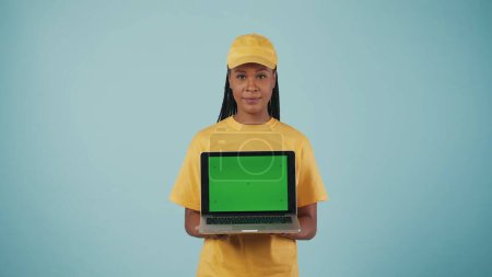 Photo for Portrait of a delivery woman in yellow cap and tshirt holding laptop in hands and looking at camera. Isolated on blue background. Advertising area, workspace mockup. - Royalty Free Image
