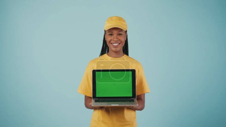 Photo for Portrait of a delivery woman in yellow cap and tshirt holding laptop and smiling at camera. Isolated on blue background. Advertising area, workspace mockup. - Royalty Free Image