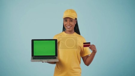 Téléchargez les photos : Portrait d'une livreuse vêtue d'une casquette et d'un t-shirt jaunes tenant un ordinateur portable et une carte de crédit, souriant à la caméra. Isolé sur fond bleu. Espace publicitaire, maquette de l'espace de travail. - en image libre de droit