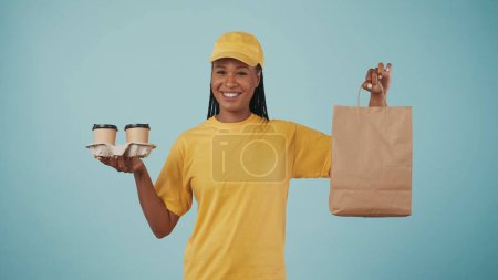 Photo for Portrait of a delivery woman in yellow cap and tshirt holding coffee cups holder and paper bag. Isolated on blue background. - Royalty Free Image