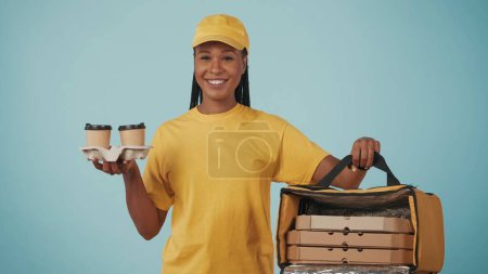 Téléchargez les photos : Portrait d'une livreuse en bonnet jaune et tshirt tenant des tasses à café et un sac à dos réfrigérateur avec pizza. Isolé sur fond bleu. - en image libre de droit