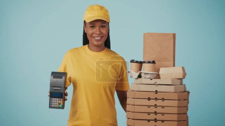 Téléchargez les photos : Portrait d'une livreuse en bonnet jaune et un t-shirt tenant terminal de paiement portable, debout à côté de tasses à café et boîtes à pizza. Isolé sur fond bleu. - en image libre de droit