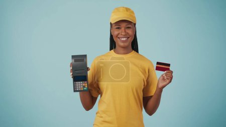Photo for Portrait of a delivery woman in yellow cap and tshirt holding portable payment terminal and bank credit card, smiling. Isolated on blue background. - Royalty Free Image