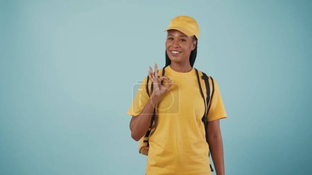 Téléchargez les photos : Portrait de la femme de livraison en casquette jaune et tshirt avec réfrigérateur sac à dos portable montre un geste correct à la caméra. Isolé sur fond bleu. - en image libre de droit