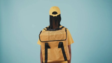 Téléchargez les photos : Portrait de la femme de livraison en bonnet jaune et tshirt avec réfrigérateur sac à dos portable, posant à l'appareil photo avec son dos. Isolé sur fond bleu. - en image libre de droit