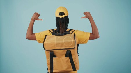 Téléchargez les photos : Portrait de la femme de livraison en casquette jaune et tshirt avec réfrigérateur sac à dos portable, montre les muscles mains en l'air. Isolé sur fond bleu. - en image libre de droit