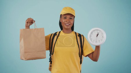 Foto de Retrato de mujer de entrega en gorra y camiseta amarilla con mochila portátil refrigerador sosteniendo bolsa de papel y reloj. Aislado sobre fondo azul. - Imagen libre de derechos