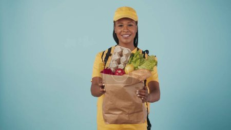 Téléchargez les photos : Portrait de la livreuse en bonnet jaune et tshirt et réfrigérateur portable sac à dos, donnant sac en papier avec des produits d'épicerie, souriant. Isolé sur fond bleu. - en image libre de droit