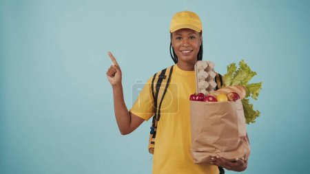 Photo for Portrait of delivery woman in yellow cap and tshirt with portable backpack refrigerator holding paper bag with grocery products. Space to insert advertisement. - Royalty Free Image
