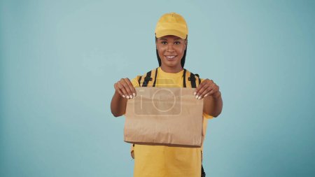 Photo for Portrait of delivery woman in yellow cap and tshirt with portable backpack refrigerator giving paper bag at the camera. Isolated on blue background. - Royalty Free Image