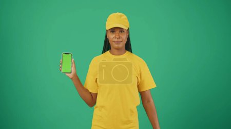 Photo for Courier service concept. Portrait of a delivery woman in yellow cap and tshirt holding smartphone. Isolated on green background. Advertising area, workspace mockup. - Royalty Free Image