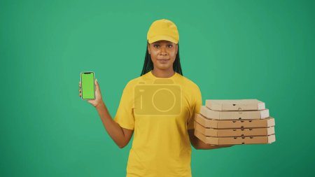 Téléchargez les photos : Portrait d'une livreuse en bonnet et t-shirt jaune tenant des boîtes à pizza et un smartphone. Isolé sur fond vert. Espace publicitaire, maquette de l'espace de travail. - en image libre de droit