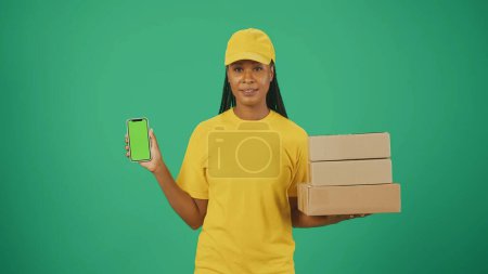 Photo for Portrait of a delivery woman in yellow cap and tshirt holding stack of boxes and smartphone. Isolated on green background. Advertising area, workspace mockup. - Royalty Free Image