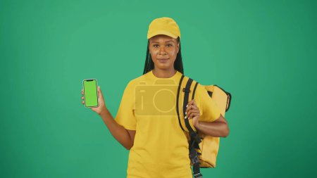 Photo for Portrait of a delivery woman in yellow cap with portable backpack refrigerator and smartphone in hand. Isolated on green background. Advertising area, workspace mockup. - Royalty Free Image