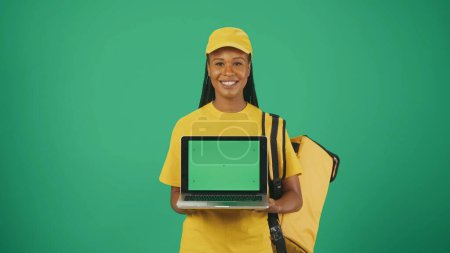 Foto de Retrato de una repartidora en gorra amarilla con mochila portátil refrigerador sosteniendo portátil y sonriendo. Área de publicidad, maqueta del espacio de trabajo. - Imagen libre de derechos