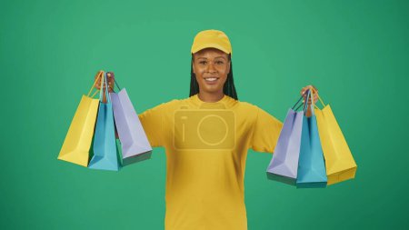 Foto de Retrato de una repartidora con gorra y camiseta amarilla sosteniendo muchas bolsas de colores y sonriendo a la cámara. Aislado sobre fondo verde. - Imagen libre de derechos