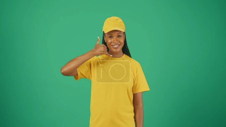 Photo for Courier service concept. Portrait of a delivery woman in yellow cap and tshirt smiling at the camera, showing calling gesture. Space to insert advertising. - Royalty Free Image