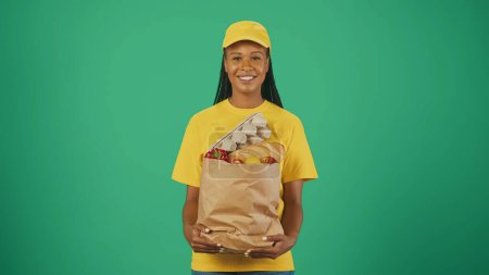 Téléchargez les photos : Portrait de la livreuse en casquette jaune et tshirt posant, souriant à la caméra avec un sac en papier rempli de produits d'épicerie, souriant. Isolé sur fond vert. - en image libre de droit