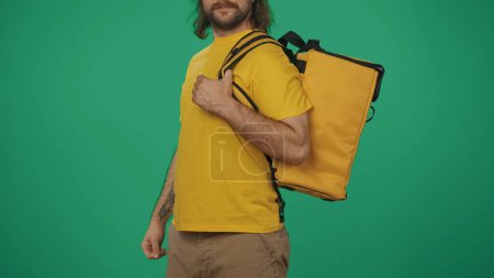 Photo for Closeup shot. Portrait of a delivery man in yellow tshirt holding portable backpack refrigerator for food products. Isolated on green background. - Royalty Free Image