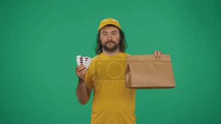 Photo for Courier service concept. Portrait of a delivery man in yellow cap and tshirt holding paper bag, pills and medicals. Isolated on green background. - Royalty Free Image