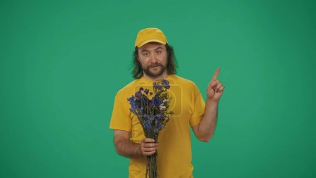 Téléchargez les photos : Portrait d'un livreur vêtu d'une casquette et d'un t-shirt jaunes tenant des fleurs et pointant vers la zone vide. Espace pour la publicité. Isolé sur fond vert. - en image libre de droit