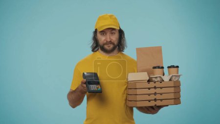 Photo for Portrait of a delivery man in yellow cap holding pizza boxes, coffee and portable payment terminal. Online payments. Isolated on blue background. - Royalty Free Image