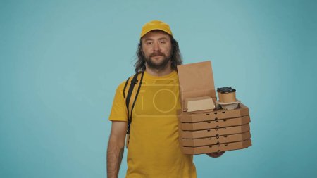 Photo for Portrait of a delivery man in yellow cap and tshirt with backpack, holding pizza, coffee and looking smiling at the camera. Isolated on blue background. - Royalty Free Image