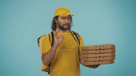 Photo for Courier service concept. Portrait of a delivery man with portable backpack refrigerator holding a stack of pizza boxes and shows ok sign. Isolated on blue background. - Royalty Free Image