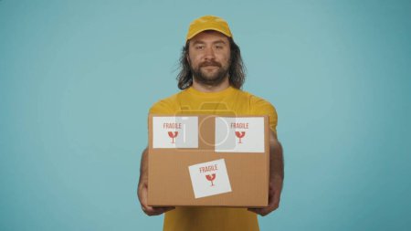Photo for Portrait of a delivery man smiling, holding a box with fragile stickers, giving it out to the camera. Isolated on blue background. - Royalty Free Image