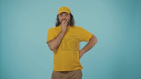 Photo for Courier service concept. Portrait of a delivery man in yellow cap and tshirt standing, posing at the camera, looking shocked and clueless. Isolated on blue background. - Royalty Free Image