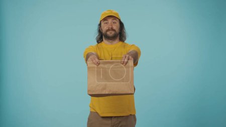 Photo for Courier concept. Portrait of a delivery man in yellow tshirt holding a paper bag in hands smiling at the camera. Isolated on blue background. - Royalty Free Image