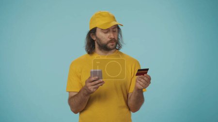 Téléchargez les photos : Portrait d'un livreur vêtu d'une casquette et d'un t-shirt jaunes tenant une carte de crédit bancaire et tapant le numéro sur son smartphone. Isolé sur fond bleu. - en image libre de droit