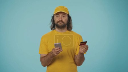 Téléchargez les photos : Portrait d'un livreur vêtu d'une casquette et d'un t-shirt jaunes tenant une carte de crédit bancaire et un téléphone intelligent, regardant la caméra. Isolé sur fond bleu. - en image libre de droit