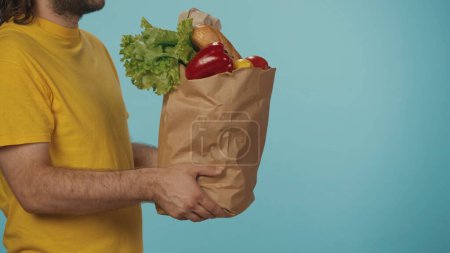 Téléchargez les photos : Gros plan. Portrait d'un livreur en tshirt jaune donnant un sac en papier avec des produits alimentaires d'épicerie au client. Isolé sur fond bleu. - en image libre de droit