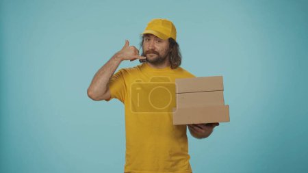 Téléchargez les photos : Concept de service de messagerie. Portrait d'un livreur en casquette jaune tenant une pile de boîtes et montrant un geste d'appel. Isolé sur fond bleu. - en image libre de droit